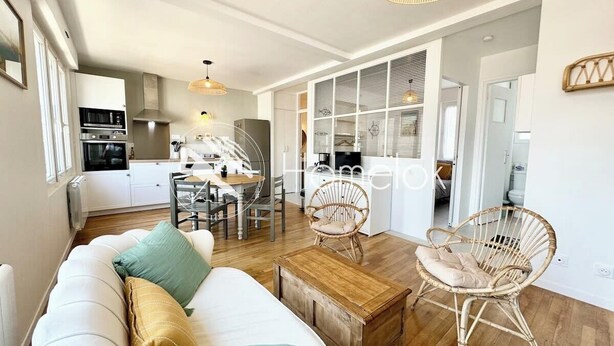 Un salon moderne avec un canapé, une table basse et des fauteuils en osier. On aperçoit à l’arrière-plan un coin repas avec une table et des chaises.