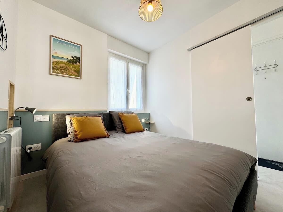 A bedroom with a bed, two pillows, a bedside table with a lamp, a radiator, a window, and a door.