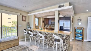 A modern kitchen with a central island, bar stools, and a dining area with a wicker sofa. - Arena De Madeira 103 - Premier (Madeira Beach)
