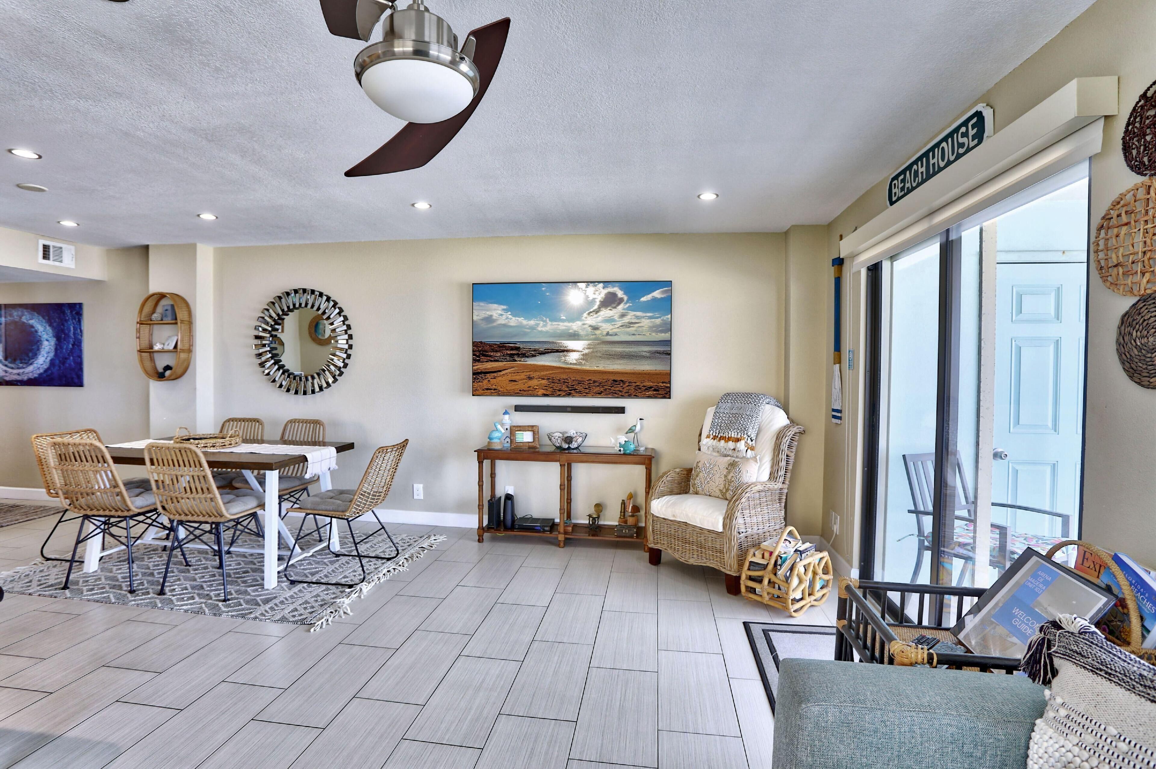 A spacious living area with a dining table, wicker chairs, a large painting of a beach, and a ceiling fan.