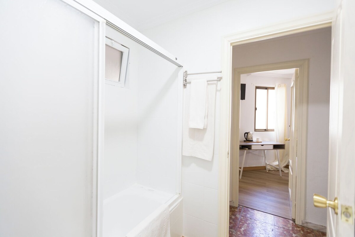 Un pasillo con una puerta blanca que da a una habitación con ventana, radiador y una pequeña mesa con tetera y taza.