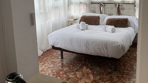 Une chambre d’hôtel avec un lit, une table de chevet avec une bouilloire, un vase de fleurs et une fenêtre avec des rideaux légers.