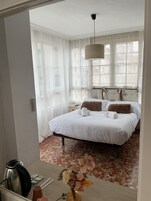 Une chambre d’hôtel avec un lit, une table de chevet avec une bouilloire, un vase de fleurs et une fenêtre avec des rideaux légers.