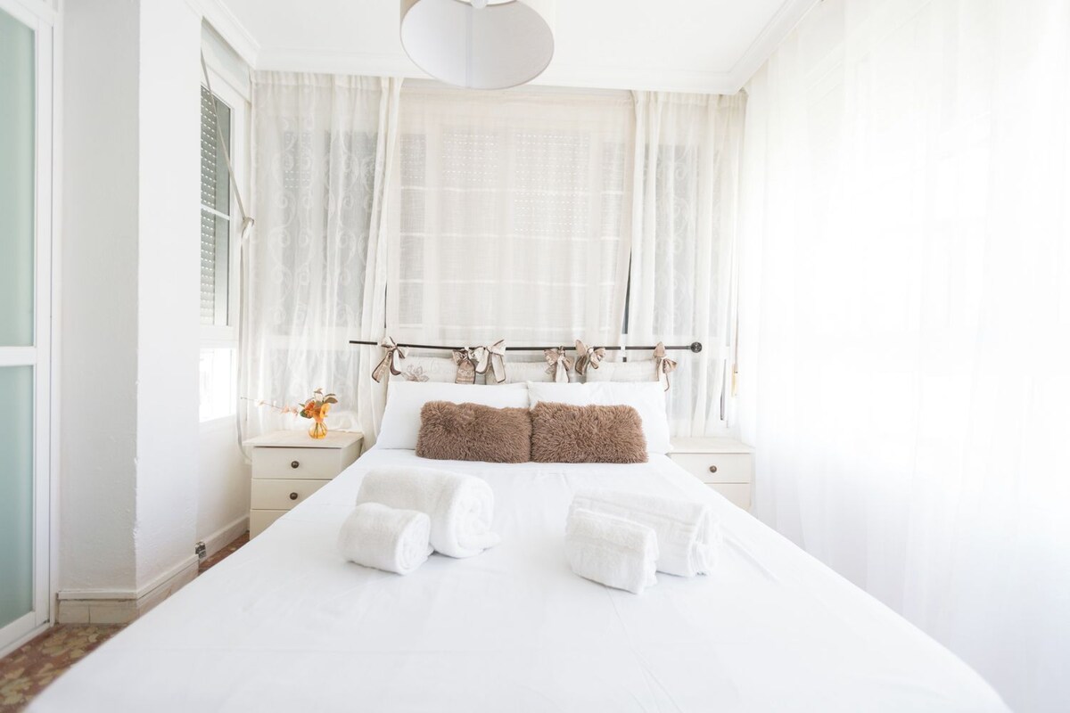 Una cama bien tendida con sábanas blancas y dos almohadas marrones, una mesita de noche con un florero y cortinas translúcidas en la ventana.