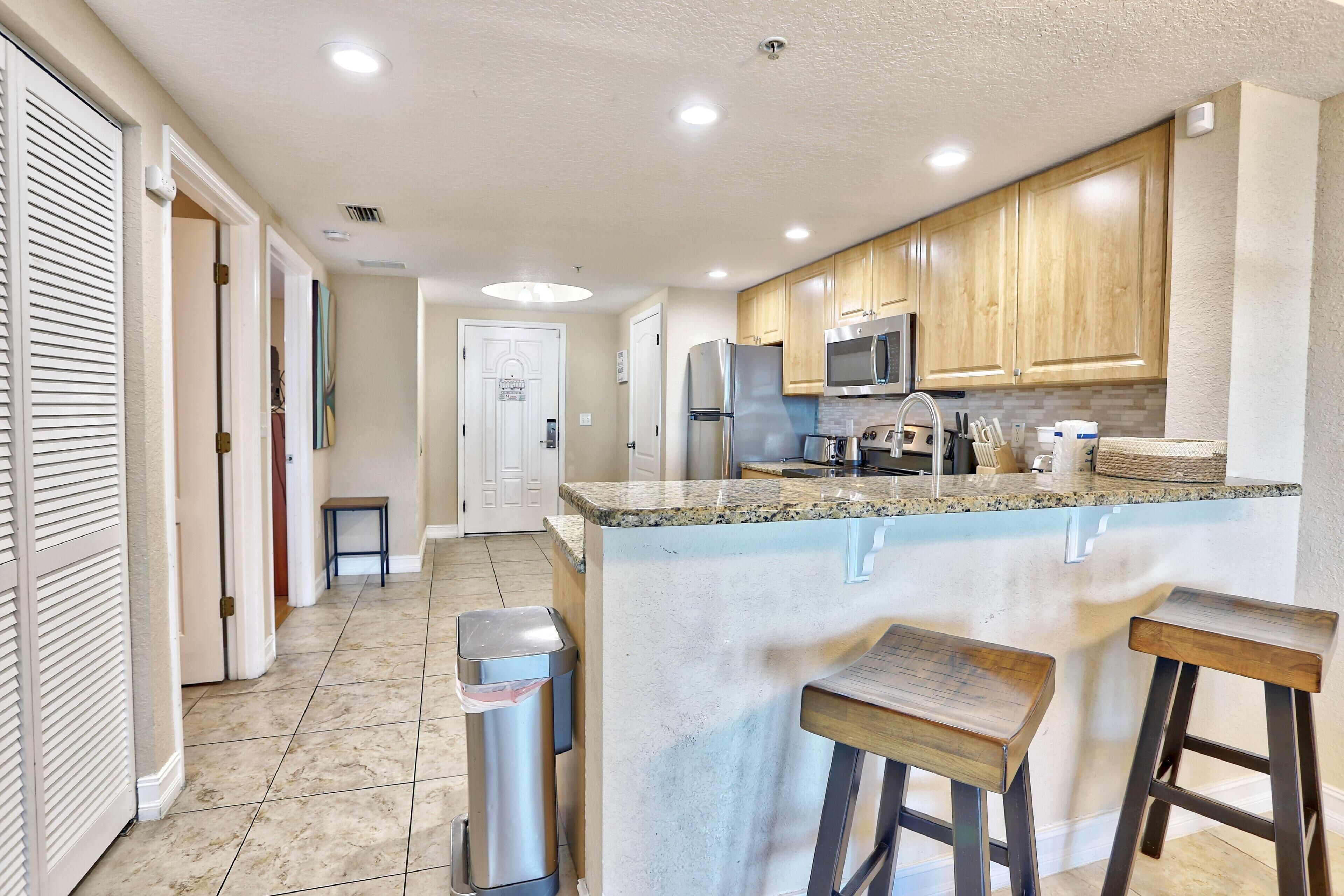 A modern kitchen with wooden cabinets, a stainless steel refrigerator, a microwave, and a granite countertop. There are two stools and a trash can.