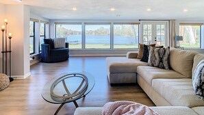 Un amplio salón con un gran ventanal con vistas a una masa de agua, un sofá modular, una mesa de centro de vidrio y una lámpara de pie.