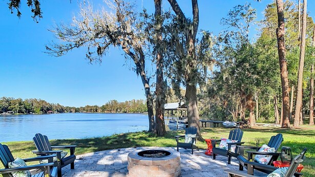 Un espacio al aire libre con sillas Adirondack y un brasero, con vista a un lago que tiene un muelle y barcos al fondo.