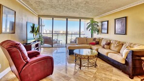 A spacious living room with a large sofa, a red armchair, a glass coffee table, and a TV on a stand. There are potted plants and a balcony with a view of the city.