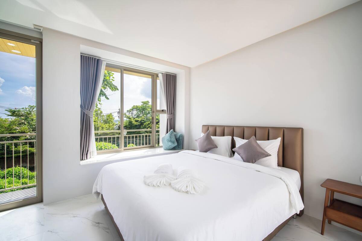 A modern hotel room with a large bed, a view of greenery, and a balcony.