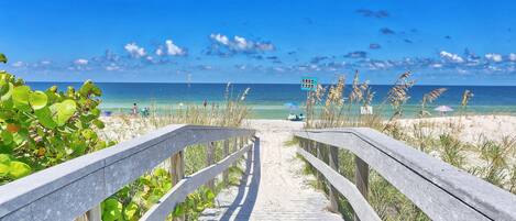 A wooden boardwalk leading to a beach with clear blue water and a sandy shore.