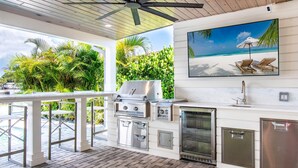 Cocina exterior con parrilla, horno y pileta, rodeada de vegetación exuberante y vista al océano.