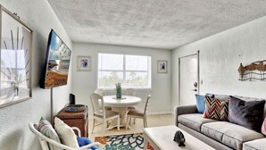 A living room with a white sofa, a dining table with chairs, a wicker side table, and a large window with blinds. - Friendly Native 204 (St. Pete Beach)