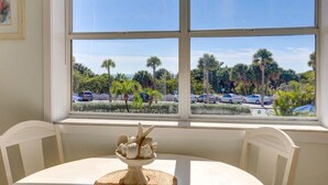 A dining area with a table and chairs, a window with a view of palm trees and parked cars. - Friendly Native 204 (St. Pete Beach)