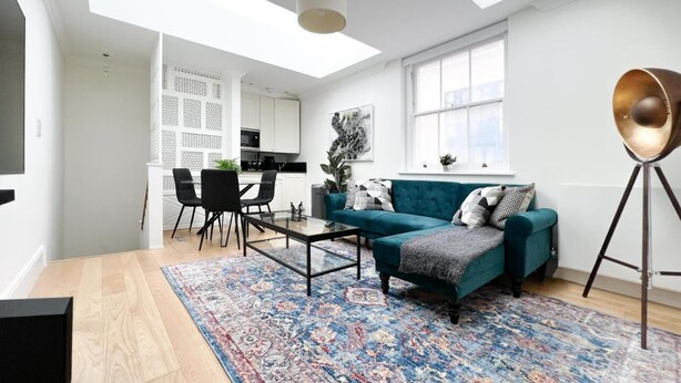 A modern living room with a teal sofa, a glass coffee table, and a patterned rug. There is a dining area with chairs and a table, and a kitchen in the background. - Museum ST Central Soho Apartment (Greater London)