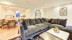 A modern living room with a grey sectional sofa, a dining table with chairs, a coffee table, and a kitchen in the background. - Prime Location Marylebone 4 Bed Designer Flat (Greater London)