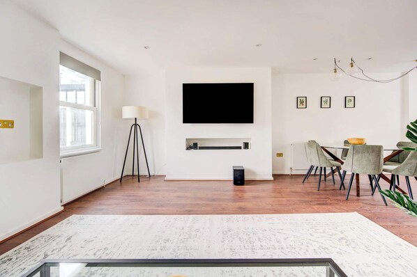 A modern living room with a flat-screen TV, a dining area with chairs, a lamp, and a large window.