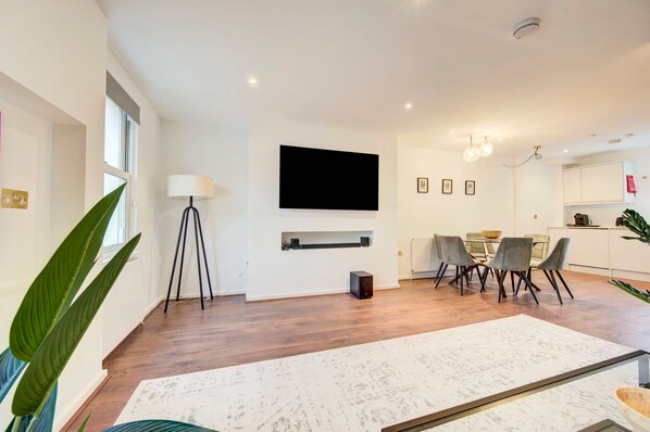 Una sala de estar moderna con televisor de pantalla plana, una mesa de comedor y sillas, una planta grande y una chimenea blanca.