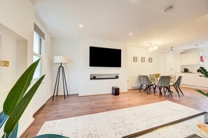 Una sala de estar moderna con televisor de pantalla plana, una mesa de comedor y sillas, una planta grande y una chimenea blanca.