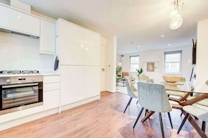 Een moderne keuken met witte kasten, een ingebouwde oven en een eethoek met een glazen tafel en stoelen.