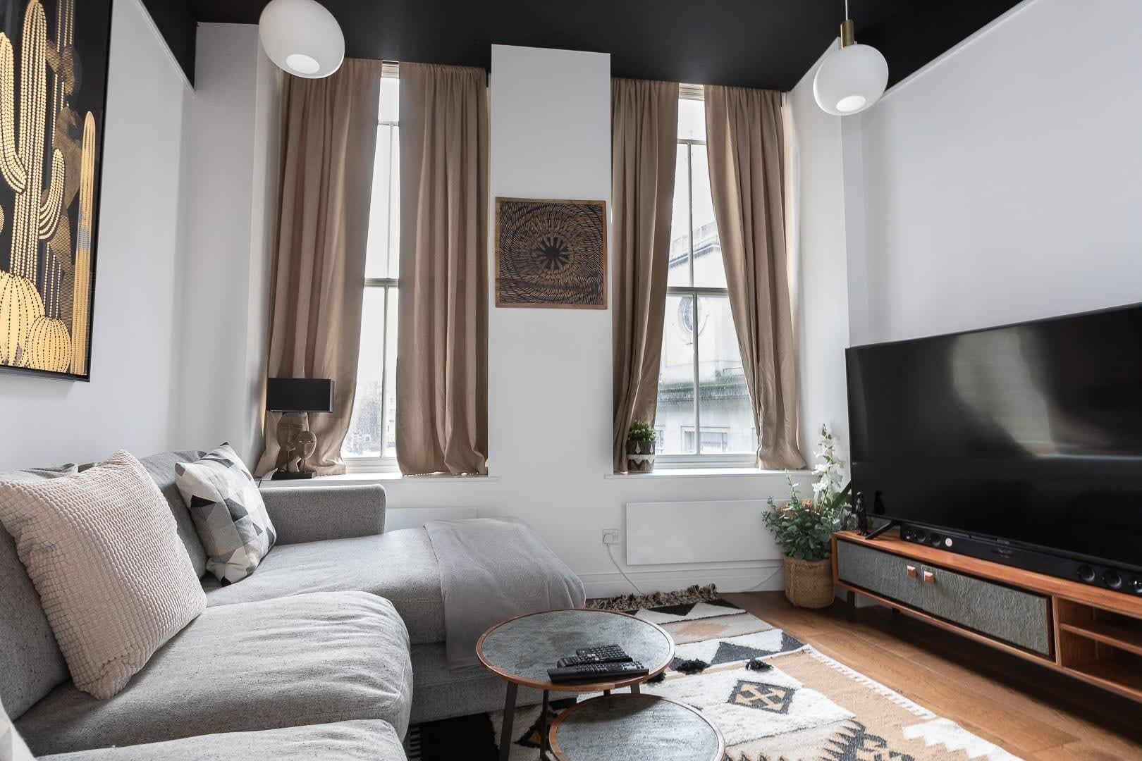 Una sala de estar moderna con un sofá gris, una mesa de centro, un televisor de pantalla plana y un amplio ventanal con cortinas.