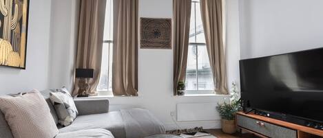 Una sala de estar moderna con un sofá gris, una mesa de centro, un televisor de pantalla plana y un amplio ventanal con cortinas.