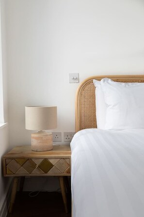 A neatly made bed with a white comforter and pillows, a bedside lamp with a beige shade, and a wooden nightstand with a geometric pattern.