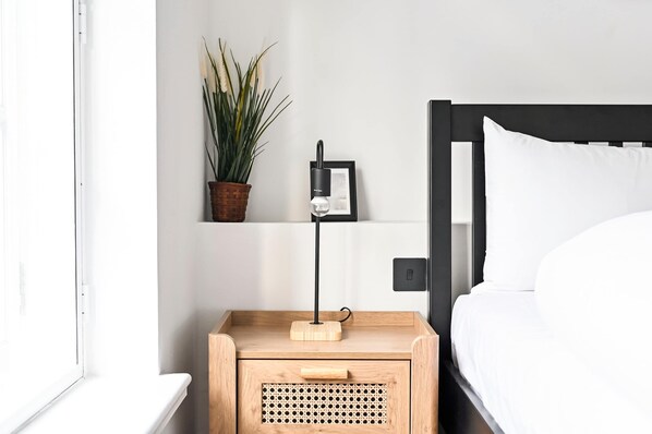 A neatly arranged bedroom with a bed, a bedside table with a lamp, a potted plant, and a framed picture on the wall.