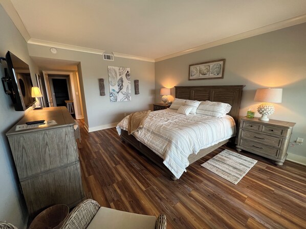 A bedroom with a large bed, wooden flooring, a dresser, a nightstand with a lamp, and a framed picture on the wall. - 438 Captain’s Walk
Oceanfront 2BR/2Bath 2nd floor condo (Hilton Head Island)