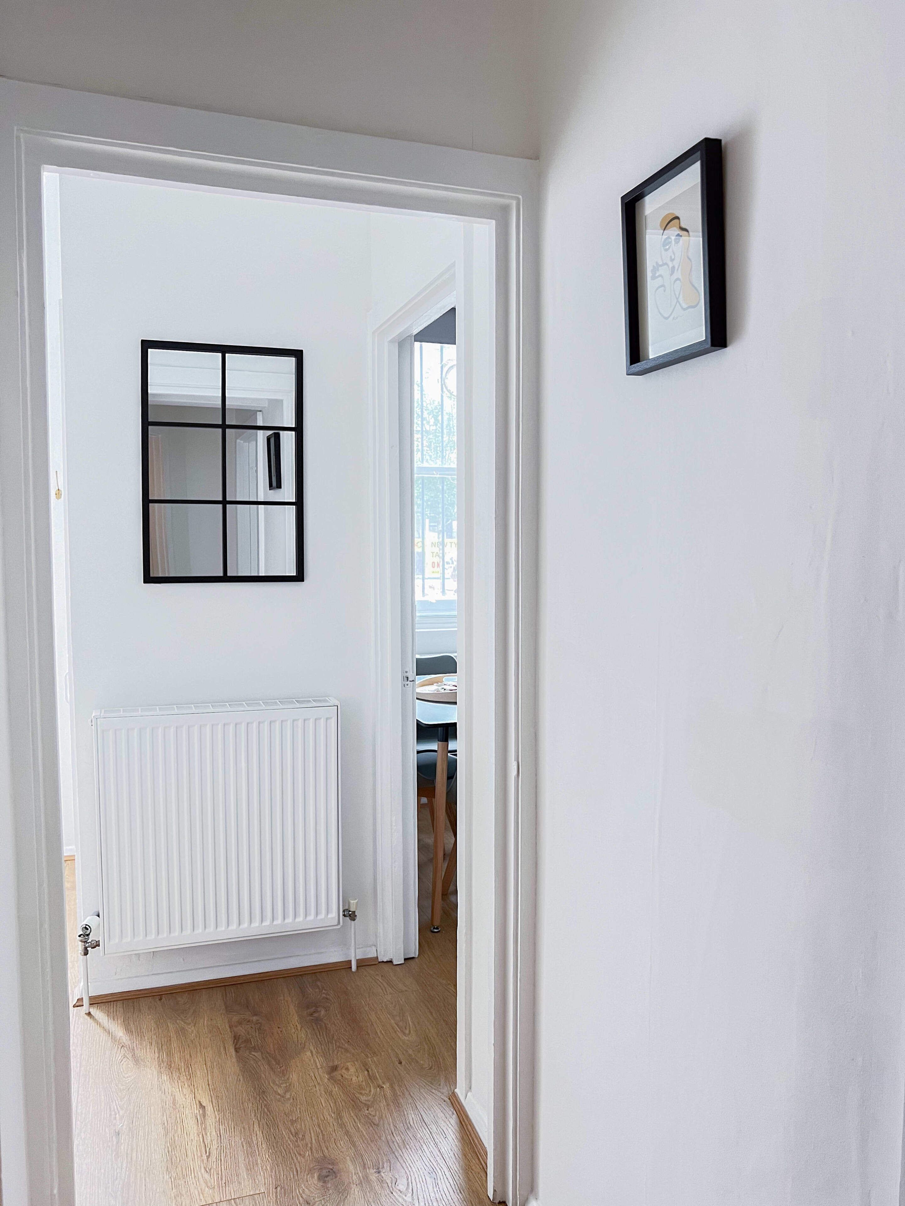Un couloir avec un radiateur blanc, un miroir à cadre noir et un tableau encadré accroché au mur.