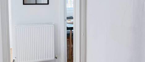 Un couloir avec un radiateur blanc, un miroir à cadre noir et un tableau encadré accroché au mur.