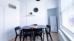 Un espace repas moderne avec une table en bois, des chaises noires, une horloge, une petite plante et un réfrigérateur blanc.