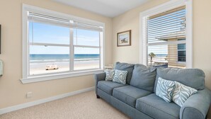 Ein Wohnzimmer mit Blick auf den Strand, ein graues Sofa mit gemusterten Kissen und ein gerahmtes Bild an der Wand.