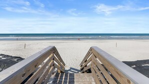 Ein hölzerner Steg führt zu einem Sandstrand, der von einem wolkenlosen, blauen Himmel überdacht wird.