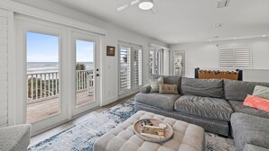 A spacious living room with a gray sofa, a wooden headboard, and a view of the ocean through large windows. - Blue Yonder Beach House (New Smyrna Beach)