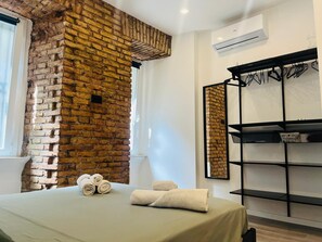 Habitación de hotel con pared de ladrillo, cama con toallas, estante con perchas y equipo de aire acondicionado.
