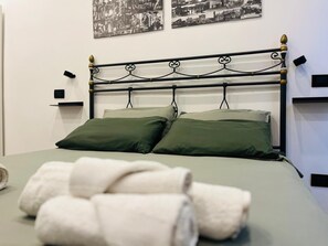 Ein Bett mit Metallgestell und zwei Kissen, ein an der Wand befestigtes Regal und ein Handtuchhalter mit Handtüchern.