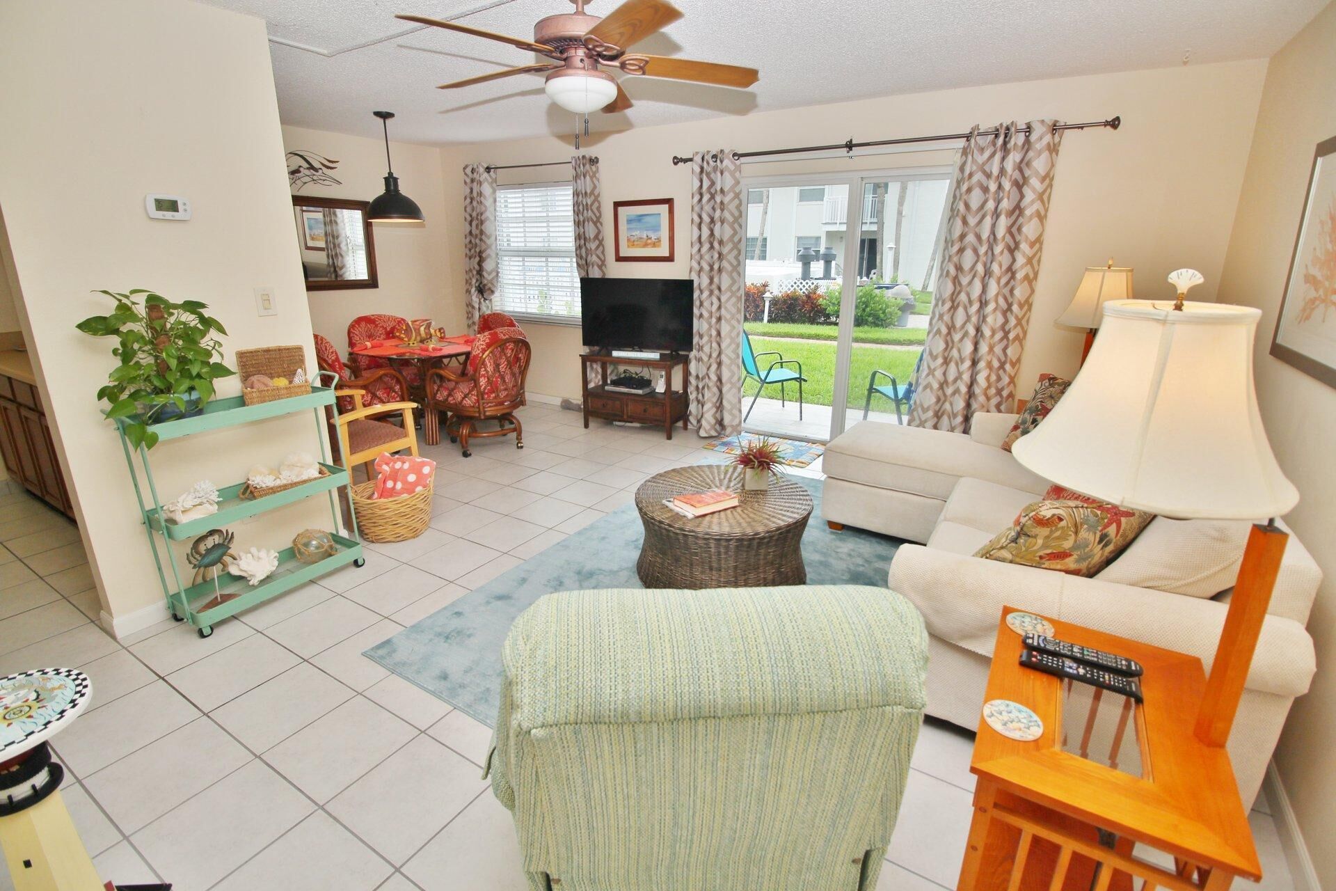 A living room with a sofa, armchair, TV, and a ceiling fan. There is a dining table with chairs and a window with curtains.