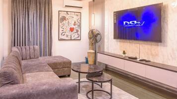 A modern living room with a sectional sofa, a small round table, a flat-screen TV mounted on the wall, and a decorative wall piece.