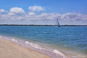 Una playa con aguas azules cristalinas, un velero a lo lejos y una costa arenosa.