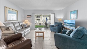 A living room with a grey leather sofa, a blue armchair, a wooden coffee table, and a TV on a stand. There is a balcony with a view of the ocean.