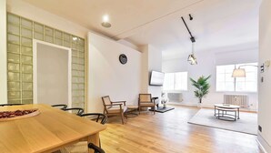 A spacious living area with a wooden floor, a dining table, and several chairs. There is a large window with blinds, a potted plant, and a radiator. - Wonderful Fitzrovia 3 Bed Apartment (Greater London)
