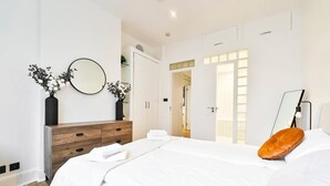 A modern bedroom with a large bed, a wooden dresser, a mirror, and a door leading to another room. - Wonderful Fitzrovia 3 Bed Apartment (Greater London)