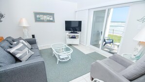 A living room with a grey sofa, a white coffee table, and a TV on a white cabinet. There is a glass door leading to a balcony with a view of the ocean.