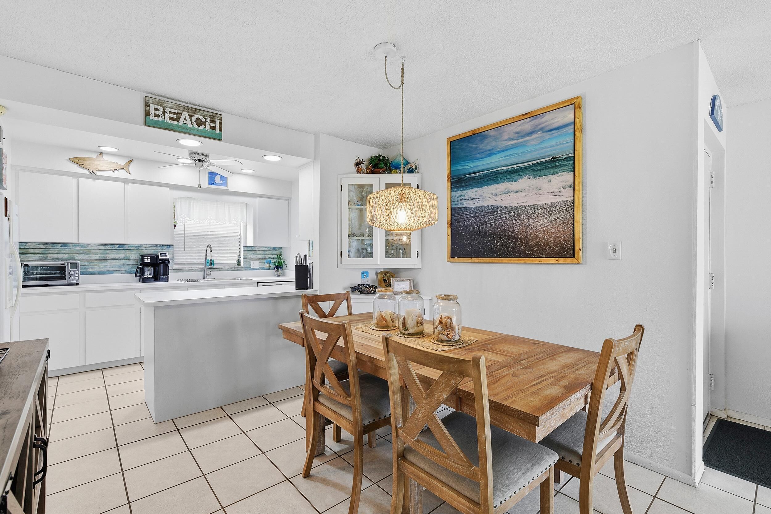 A modern kitchen with white cabinets, a wooden dining table, and a beach-themed sign.