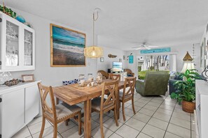 Un coin repas avec une table et des chaises en bois, un tableau représentant une plage et un ventilateur de plafond.