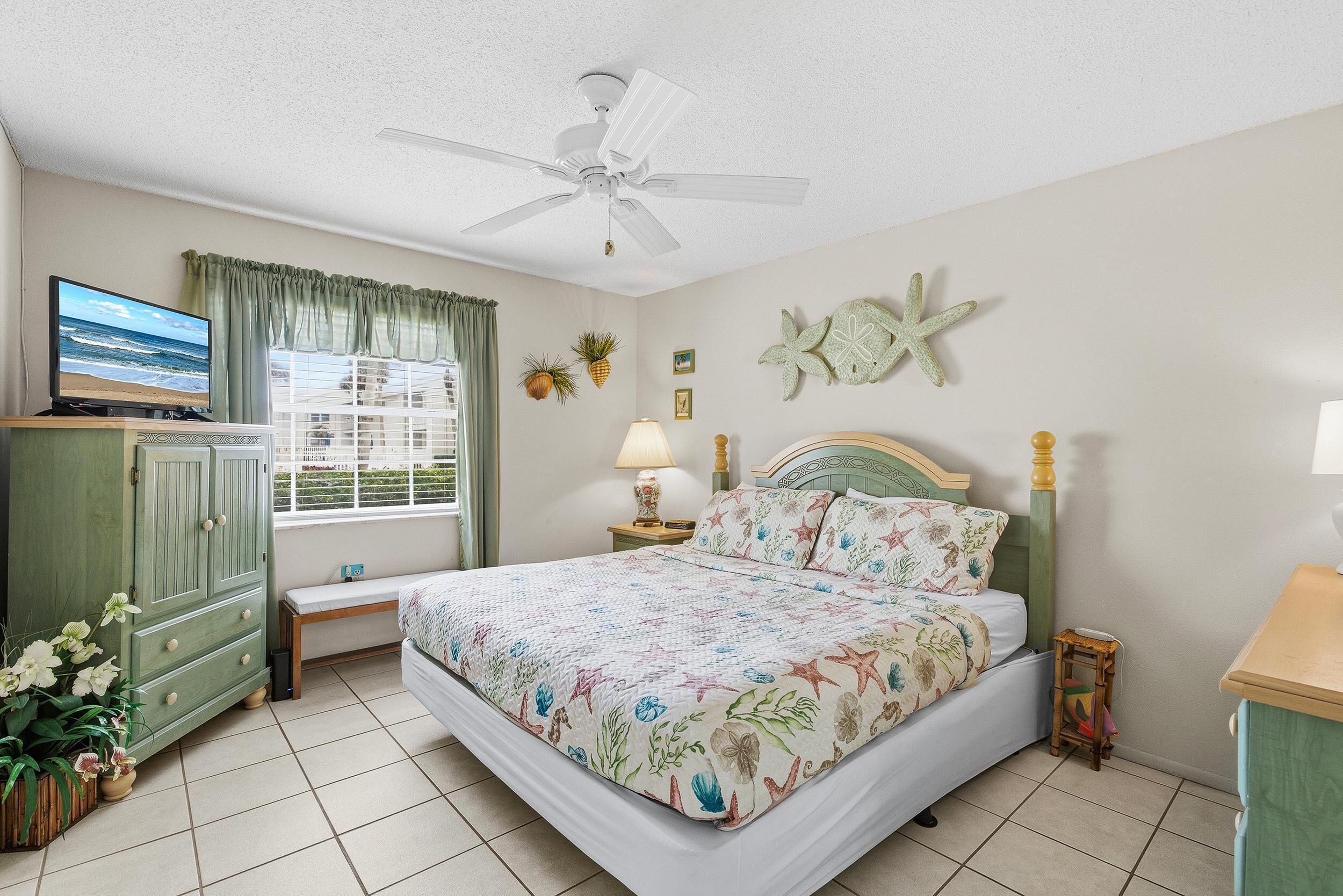 A bedroom with a bed, a ceiling fan, a TV, a window with curtains, and a wall with a starfish decoration.