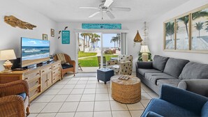 Sala de estar com televisão, móveis de vime, sofá cinza e vista para o exterior.