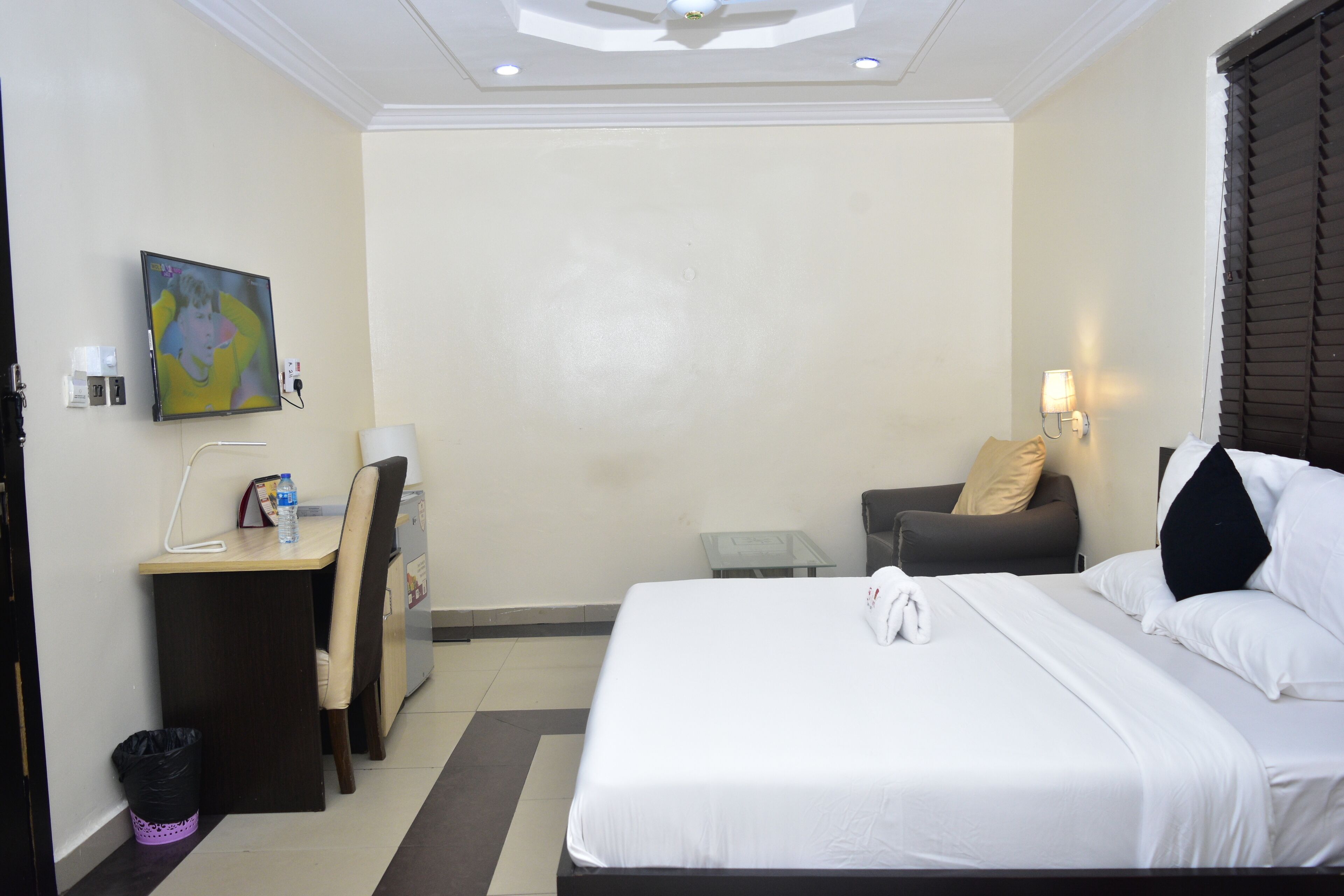 A hotel room with a bed, a desk with a chair, a small table, a trash bin, and a wall-mounted TV.
