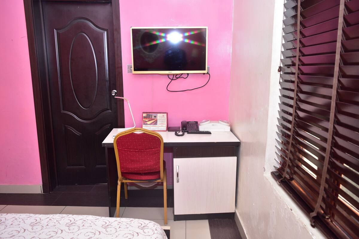 Una habitación con una cama, un escritorio con silla, un televisor en la pared, una puerta y una ventana con persianas.