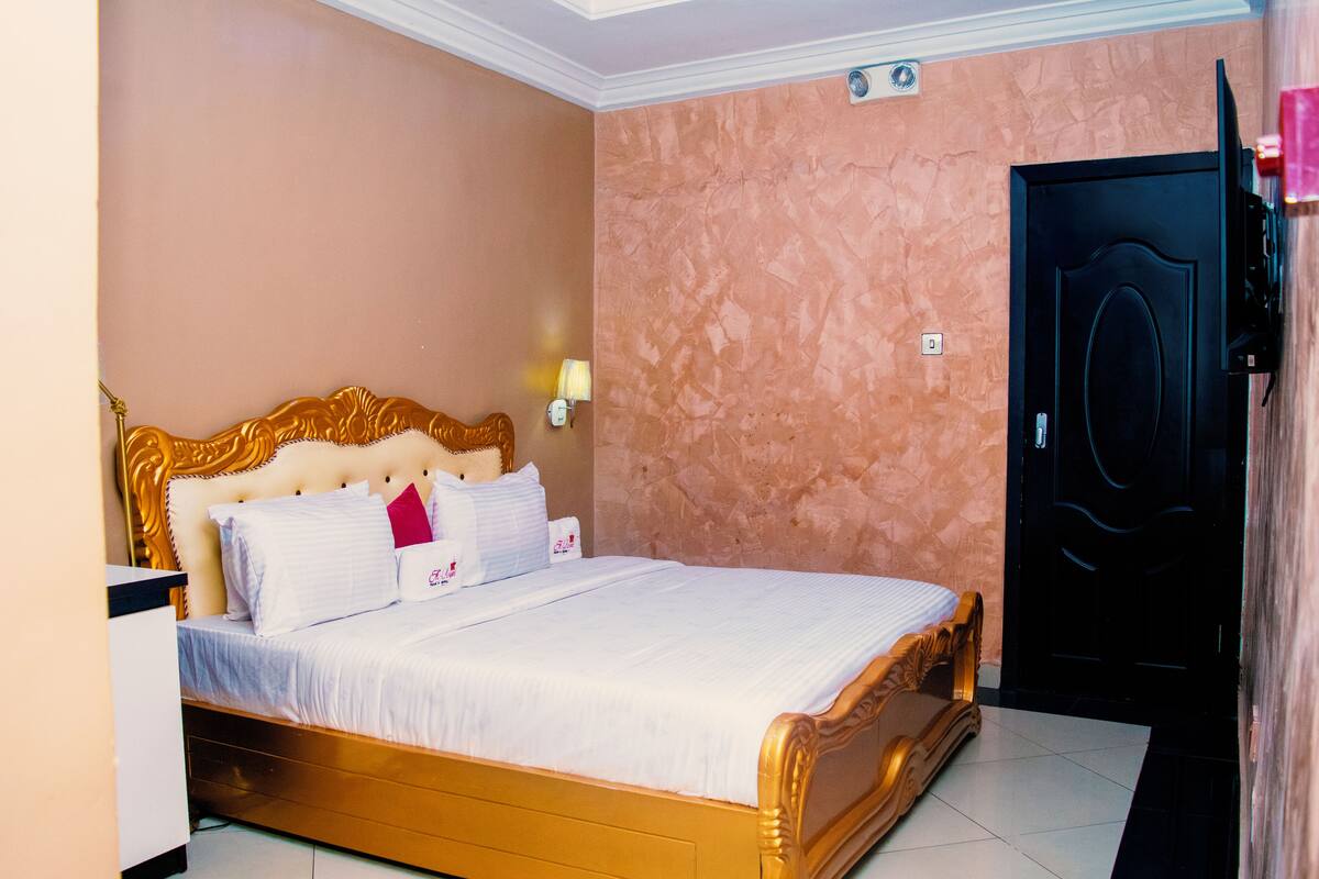 Habitación de hotel con una cama grande, cabecera de madera, mesita de noche, lámpara y una puerta.